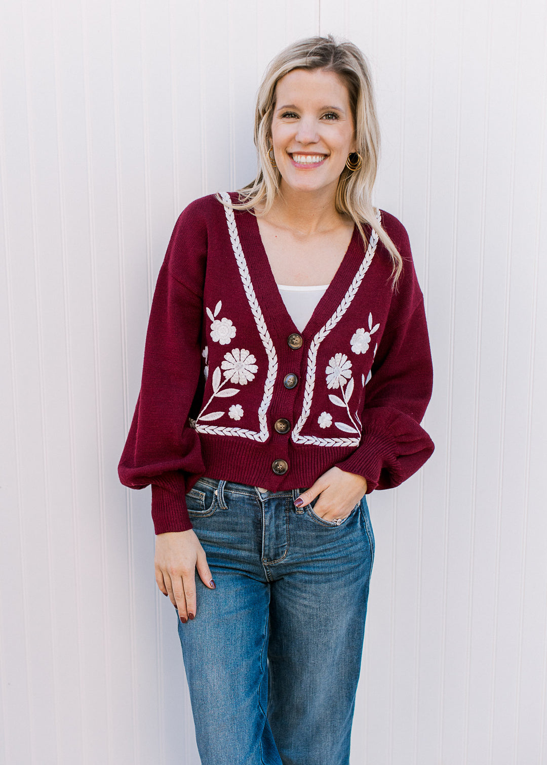 Model wearing jeans and a wine sweater with white embroidery, button front and long sleeves.