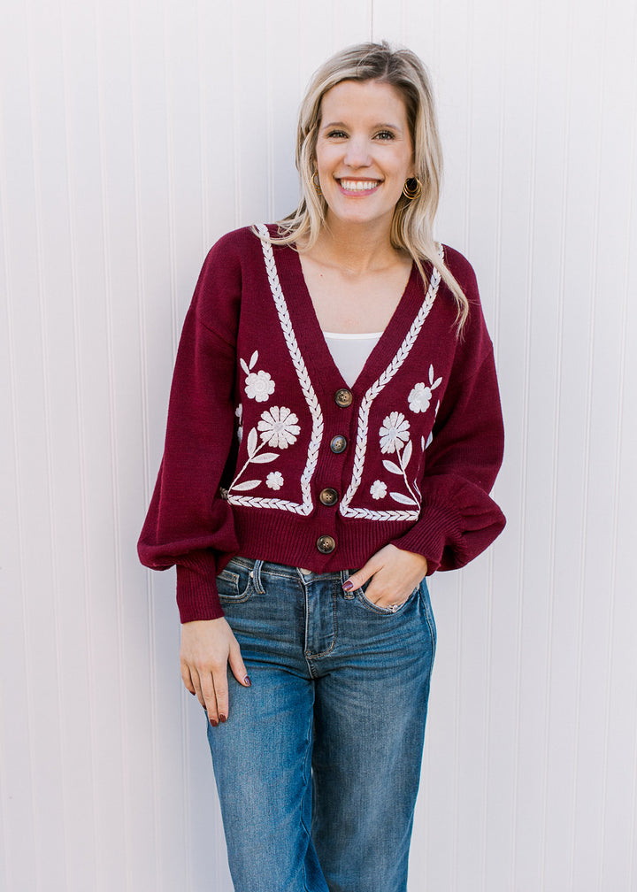 Model wearing jeans and a wine sweater with white embroidery, button front and long sleeves.