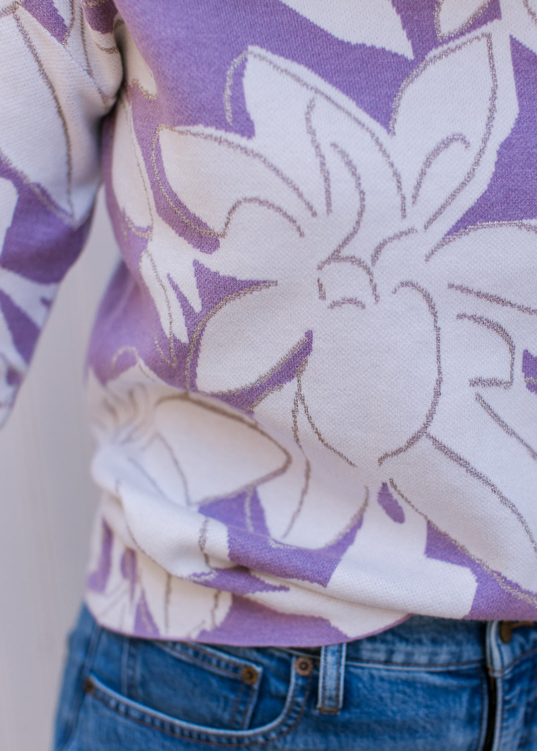 Close up of metallic detail and cream flowers on a lavender sweater with long sleeves.