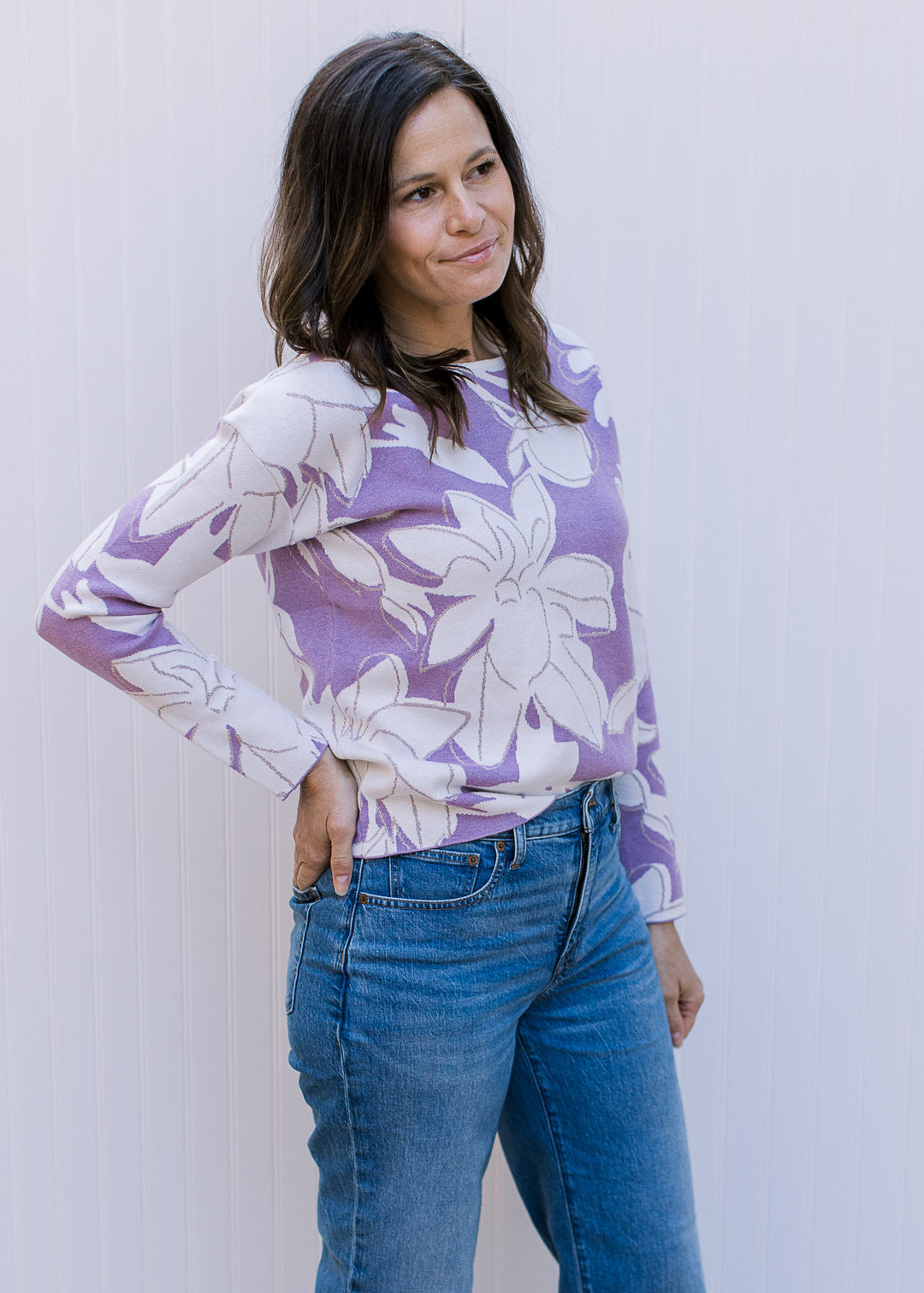 Model wearing jeans and a lavender long sleeve sweater with cream flowers and metallic details.