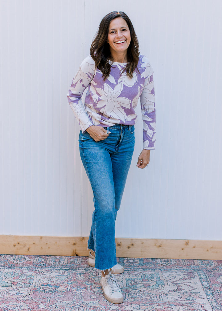 Model wearing jeans, sneakers and a lavender sweater with cream flowers and metallic details.