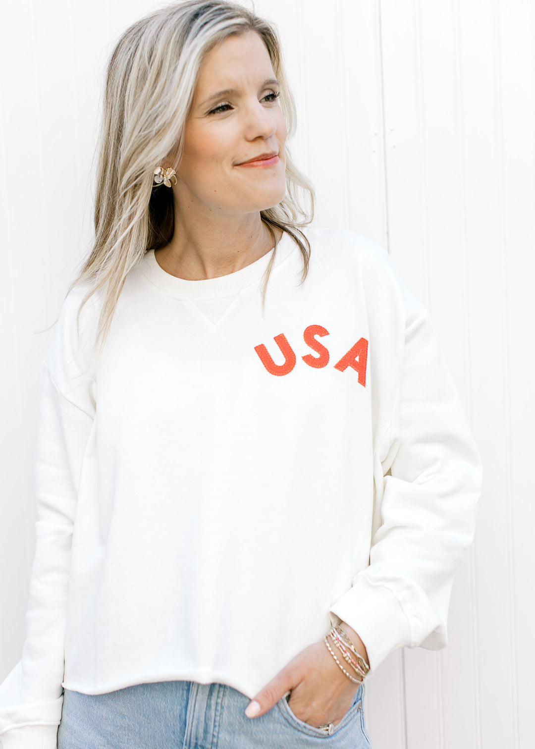 Model wearing a slightly cropped white sweatshirt with a fabric USA patch and ribbed neck.