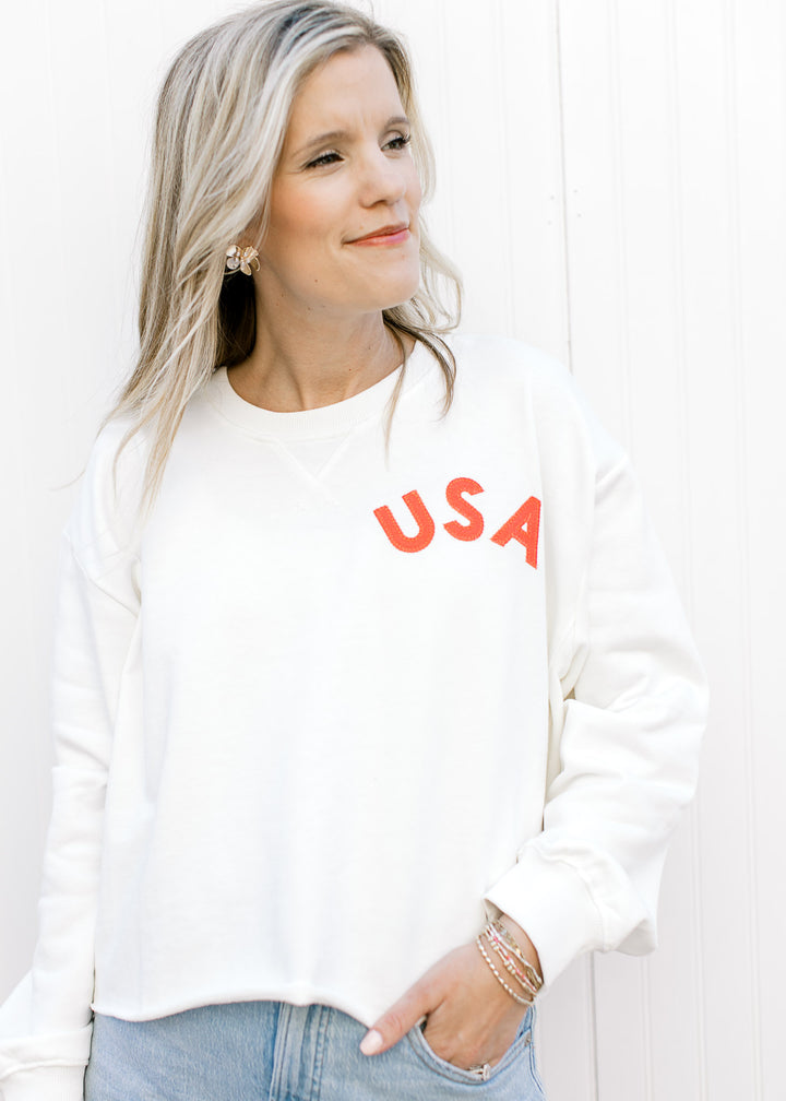 Model wearing a slightly cropped white sweatshirt with a fabric USA patch and ribbed neck.