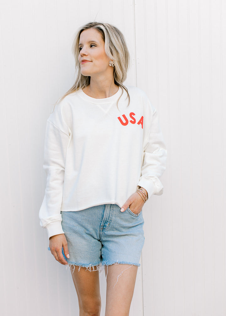 Model wearing jean shorts and a slightly cropped white sweatshirt with a fabric USA patch.