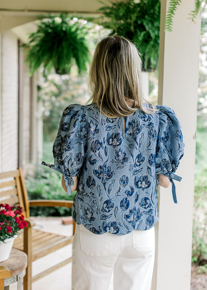 Back view of Model wearing a soft blue top with navy flowers and bubble short sleeves with a tie.