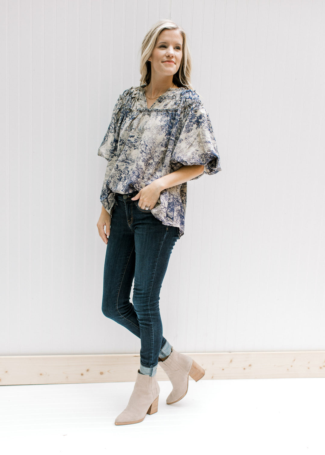 Model wearing jeans and booties with a tan top with navy toile print and bubble short sleeves.