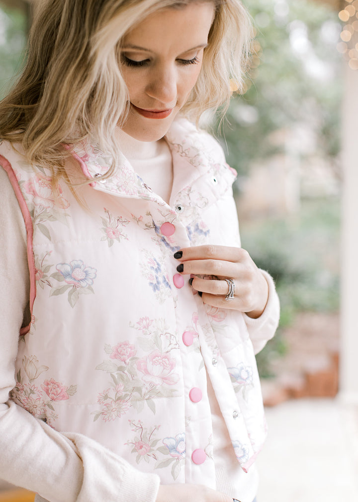 Model wearing a pale pink vest with  soft colored flowers, mock neck, pockets and snap closure.