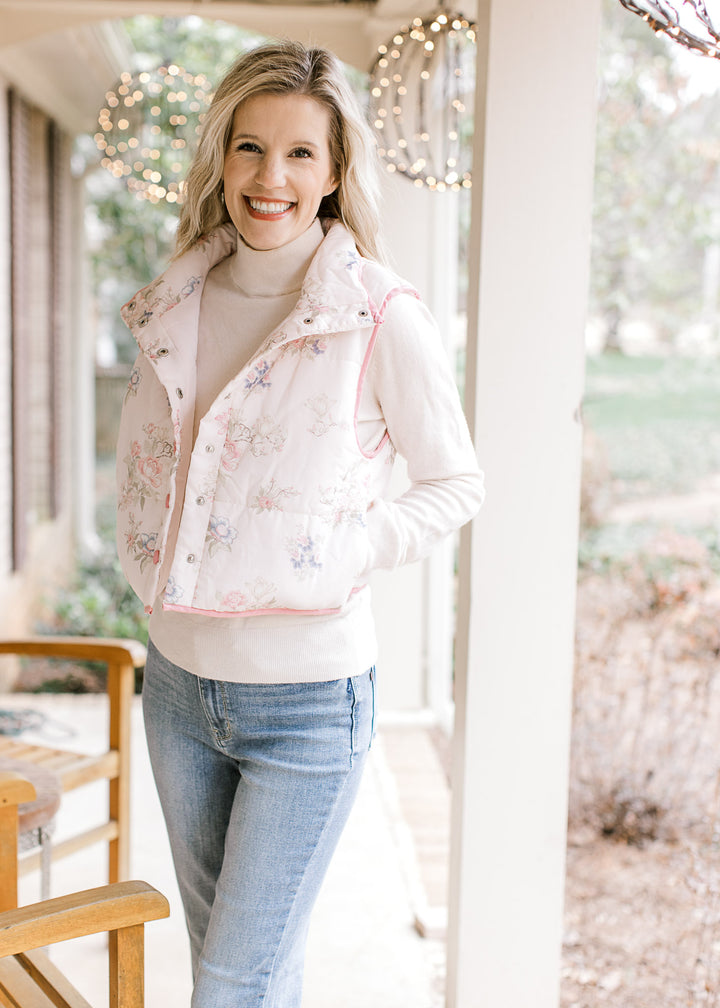 Model wearing a pale pink puffer vest with  flowers, mock neck, pockets and snap closure.