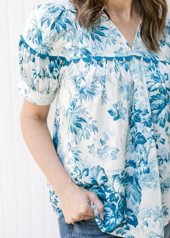 Close up of scallop detail along yolk of a cream v-neck top with teal flowers.