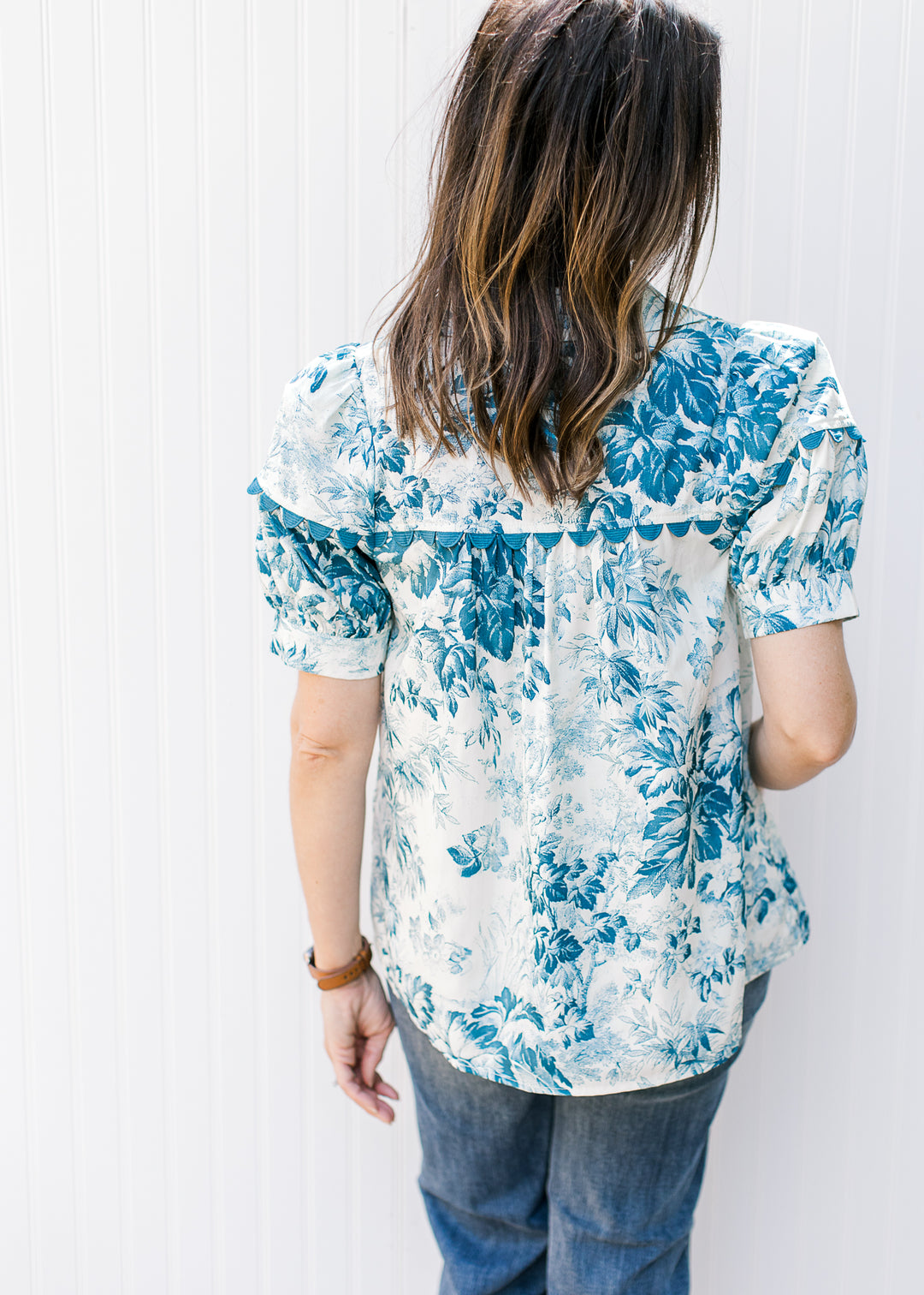 Back view of Model wearing a cream top with teal flowers, short sleeves and scallop detail