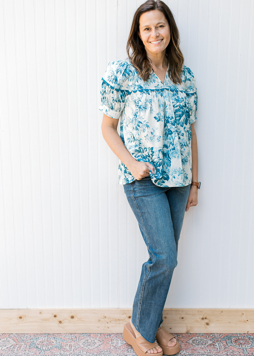 Model wearing jeans with a cream top with teal flowers, short sleeves and scallop detail
