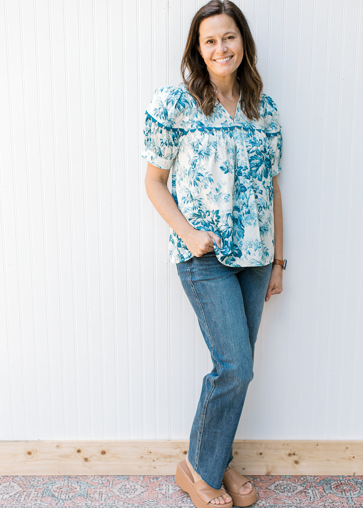 Model wearing jeans with a cream top with teal flowers, short sleeves and scallop detail