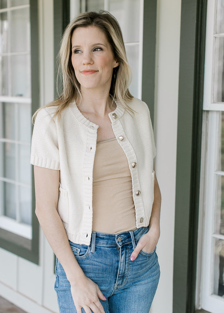Model wearing a cream short sleeve sweater with gold buttons and patch pockets.