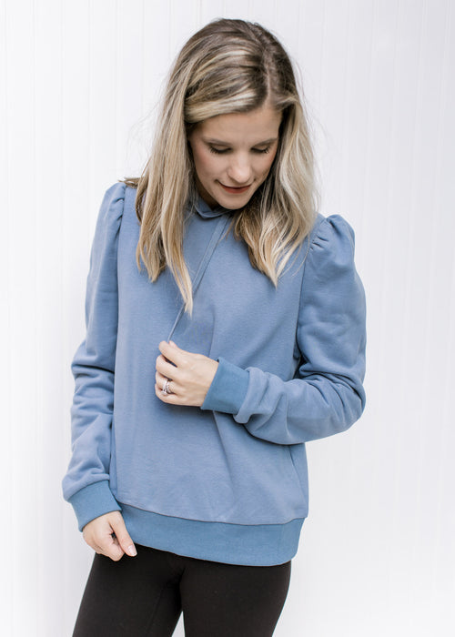 Model wearing a dusty blue pullover with puff shoulders, long sleeves and a hoodie. 