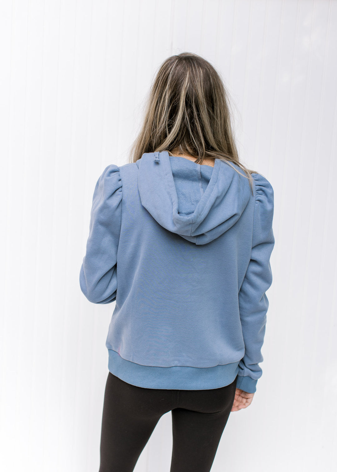 Back view of Model wearing a dusty blue pullover with puff shoulders, long sleeves and a hoodie.