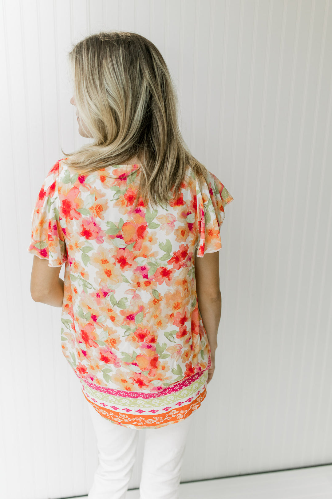 Back view of Model wearing a white top with red, orange and green floral print with short sleeves.