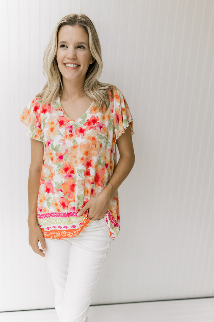 Model wearing white jeans with a white v-neck top with red, orange and green floral print.