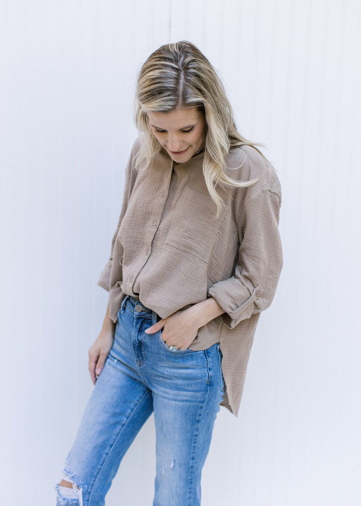 Model wearing a taupe button up top with a raw hemline, a cotton gauze material and long sleeves.
