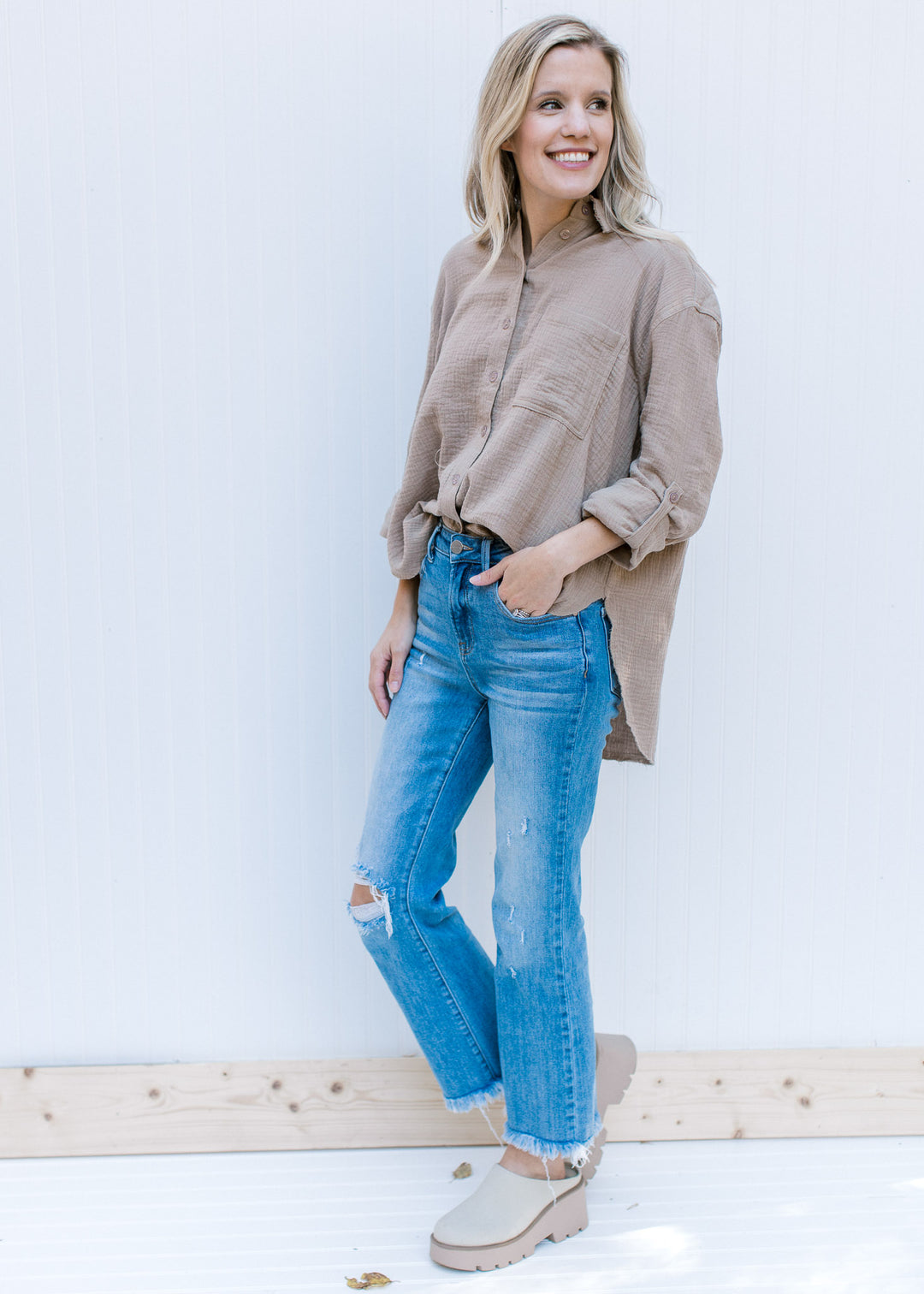 Model wearing jeans, mules and a taupe button up top with a raw hemline, a cotton gauze material.