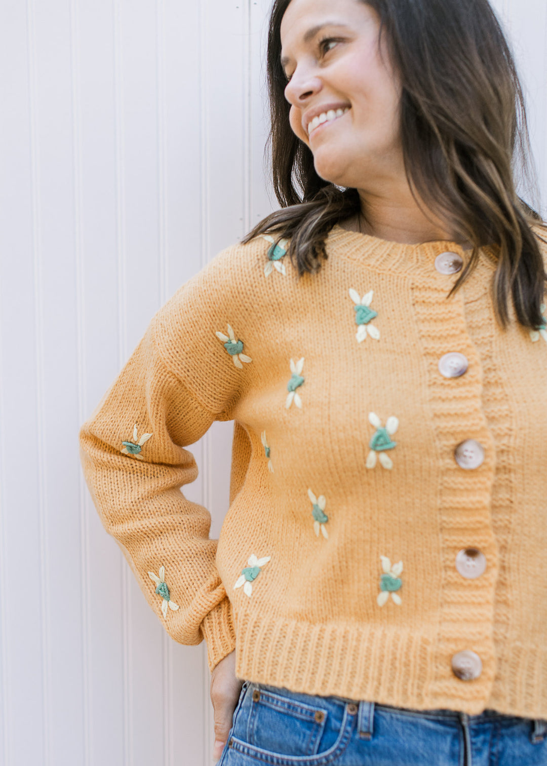 Model wearing a mustard colored button up sweater with yellow and green stitched flowers.