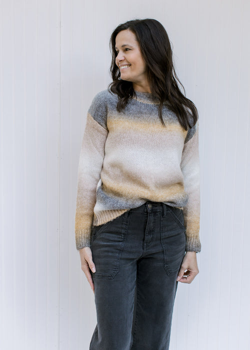 Model wearing jeans with a long sleeve gray, cream and mustard ombre sweater with a ribbed detail. 