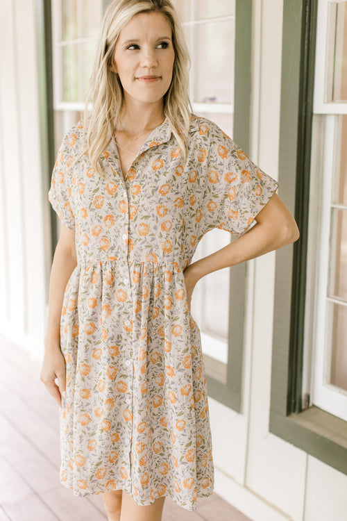 Model wearing a tan button up dress with orange flowers, short sleeves and a collar. 