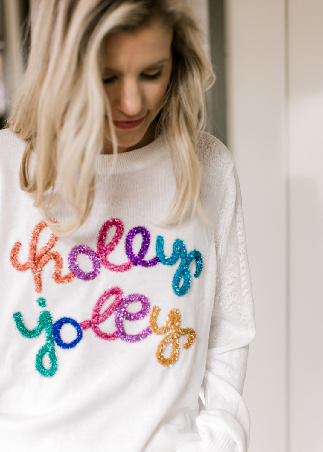 Close up of the words holly jolly in multicolored tinsel across a white sweater with long sleeves.