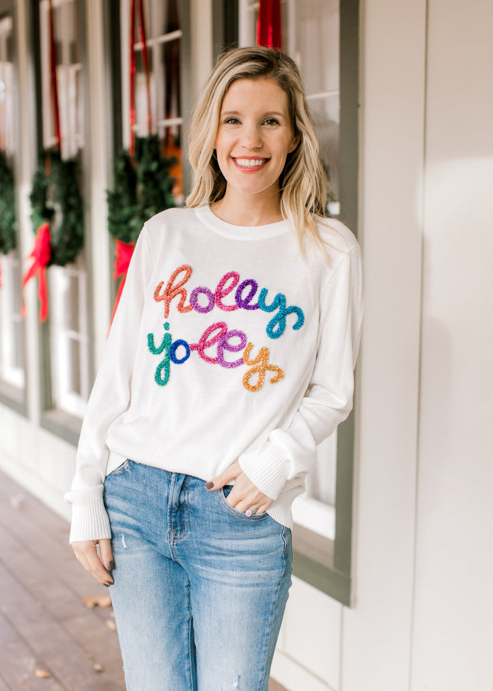 Model wearing jeans with a white sweater with holly jolly across the front in colored tinsel.