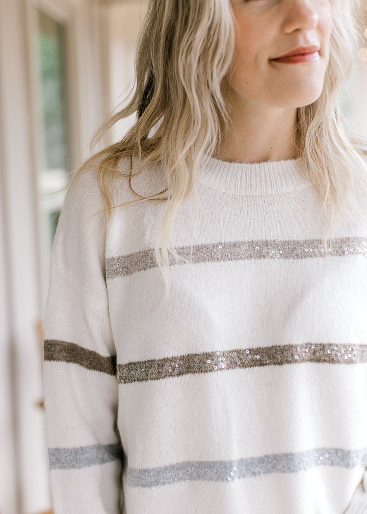Model wearing a cream long sleeve sweater with gold, champagne and silver stripes with sequins.