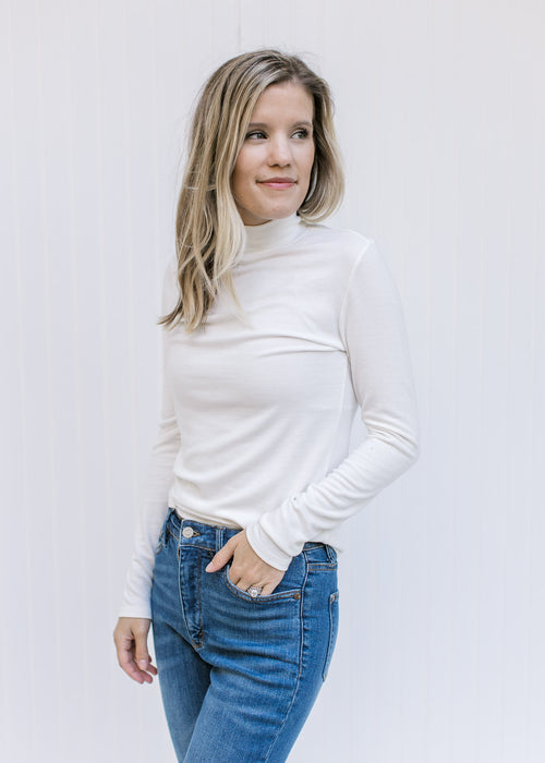 Model wearing jeans with an ivory form fitted top with a mock neckline and long sleeves. 