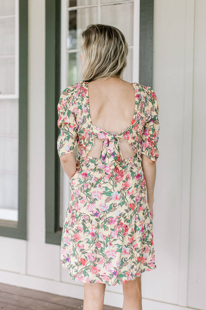 Back view of a model wearing a yellow dress with pink and green floral and a tie back closure.
