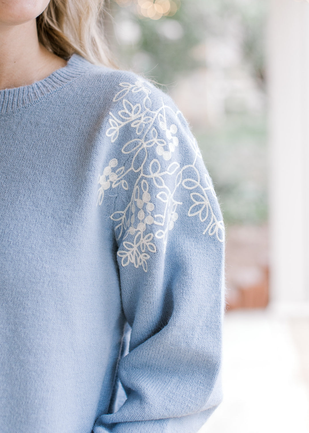 Close up of cream embroidered flowers at the shoulder of a dusty blue long sleeve sweater.