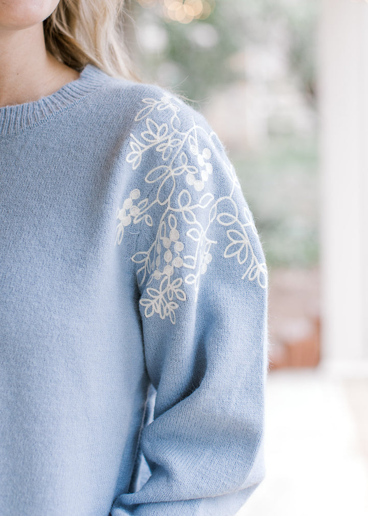 Close up of cream embroidered flowers at the shoulder of a dusty blue long sleeve sweater.