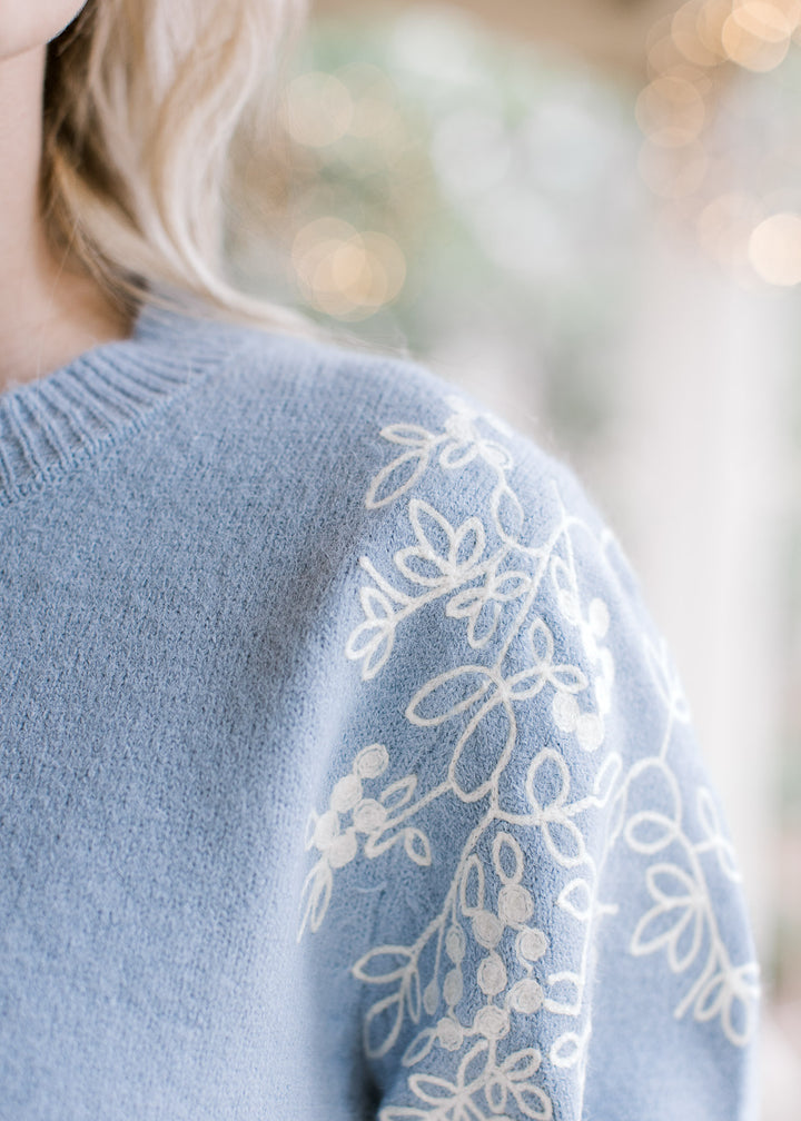 Detailed view of cream embroidery at the shoulder of a dusty blue long sleeve sweater.