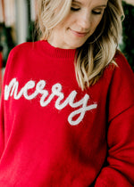 Close up of white merry in scripted letters across a long sleeve, round neck red sweater. 