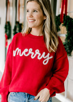 Model wearing a red long sleeve sweater with merry scripted in white across the front. 
