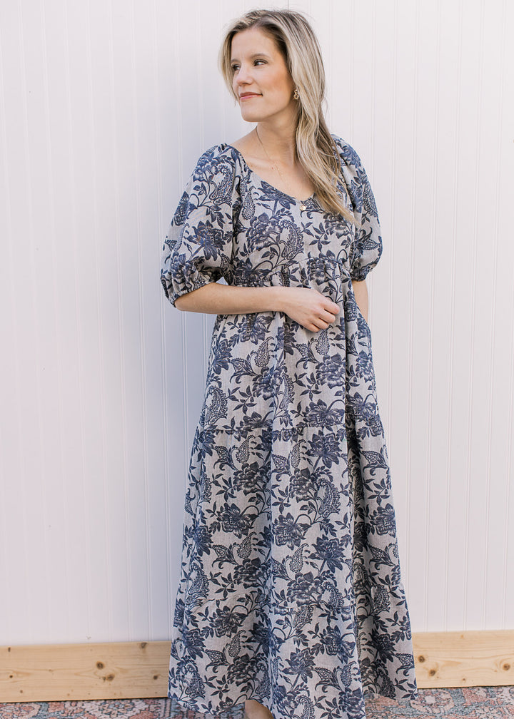 Model wearing a gray, v-neck maxi with navy floral, short puff sleeves and pockets.