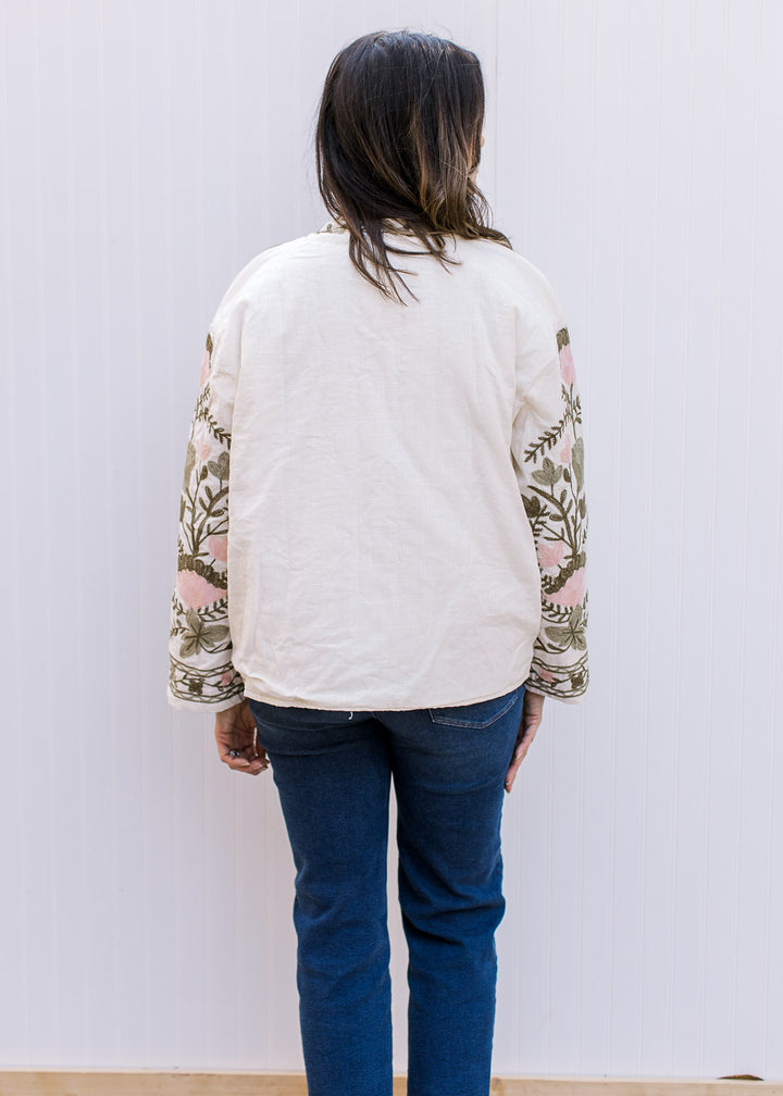 Back view of Model wearing a cream open front jacket with embroidered flowers and long sleeves.