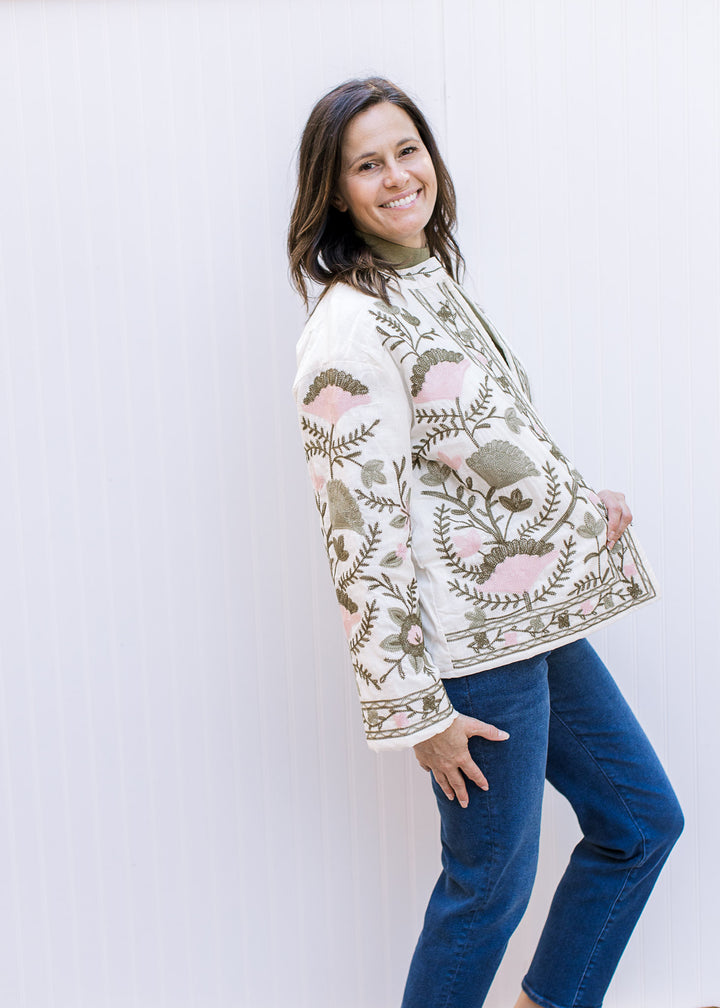 Model wearing jeans and a cream open front jacket with pink and green flowers and long sleeves.