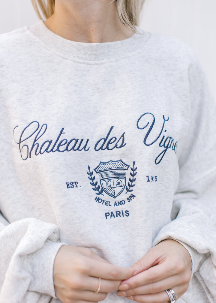 Model wearing a gray sweatshirt with a navy Paris embroidery, long sleeves and a crew neckline.