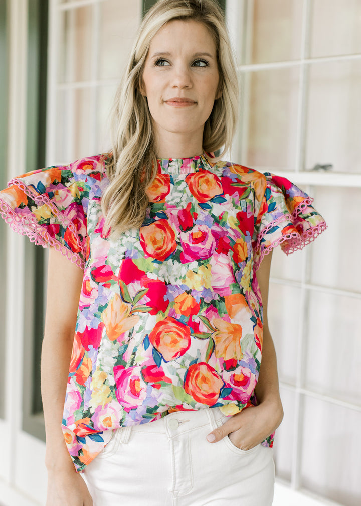 Model wearing a bright floral top with flutter short sleeves with lace and ruffle neck.