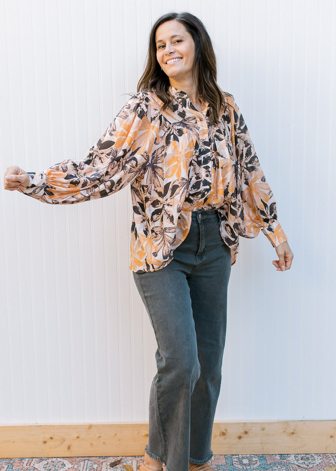 Model wearing black jeans and a cream top with brown and orange flowers and long sleeves.