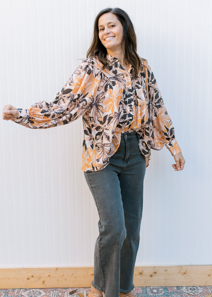 Model wearing black jeans and a cream top with brown and orange flowers and long sleeves.