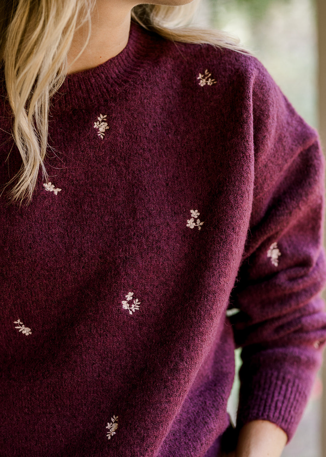 Model wearing a long sleeve plum sweater with cream embroidered flowers and a round neck.