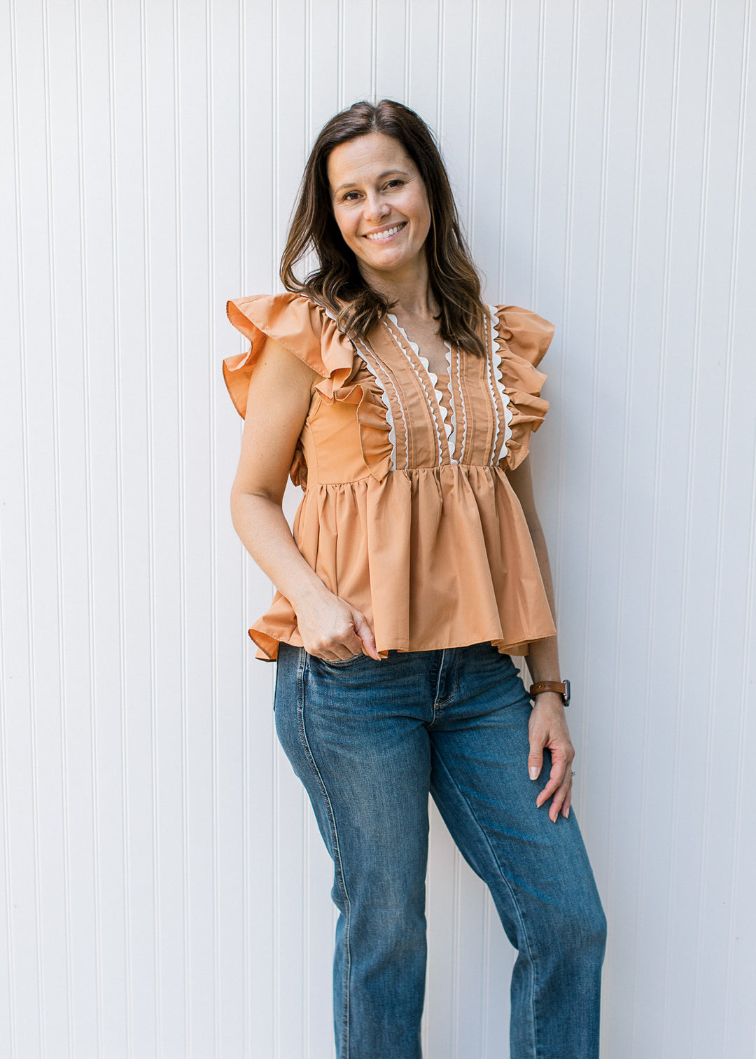 Model wearing jeans with a dusty orange top with ruffle cap sleeve and scallop white detail.