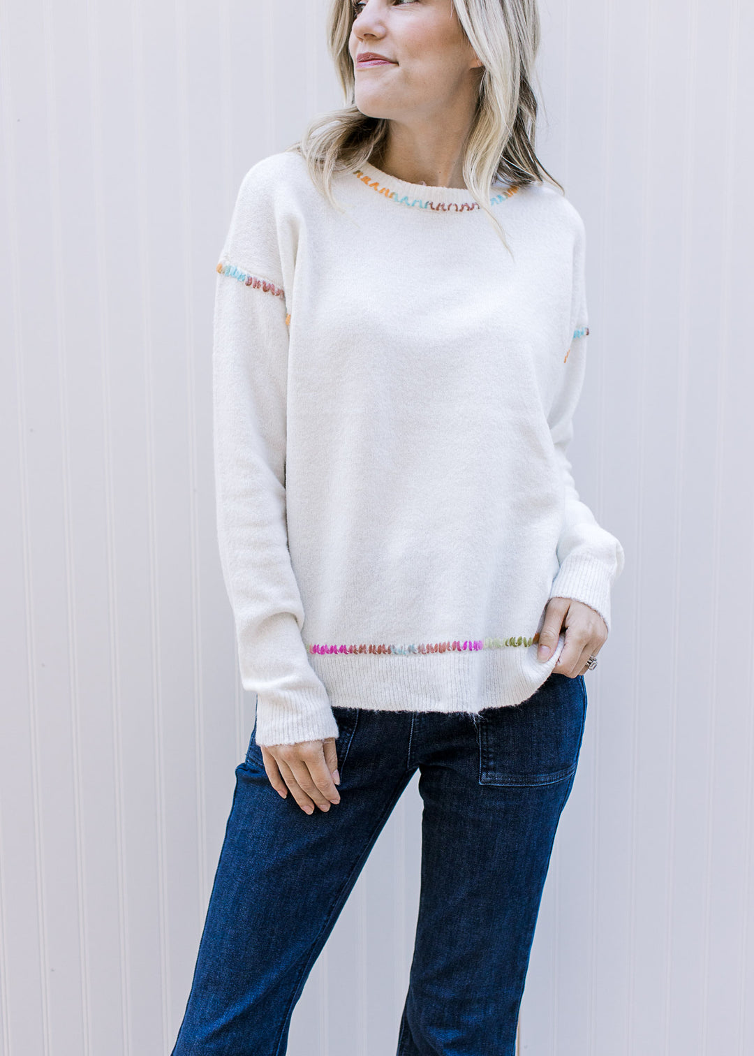 Model wearing an off white sweater with rainbow stitching at the collar, shoulders, and waist.