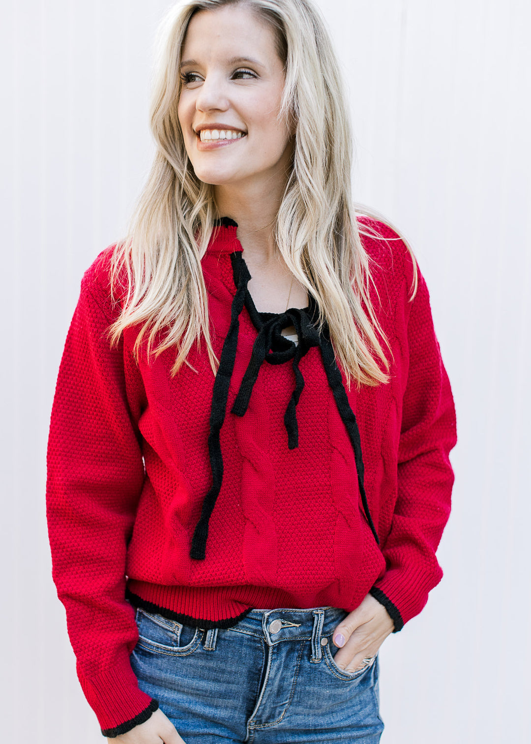 Model wearing a red sweater with black tie bow at v-neck, cable knit detail and long sleeves.