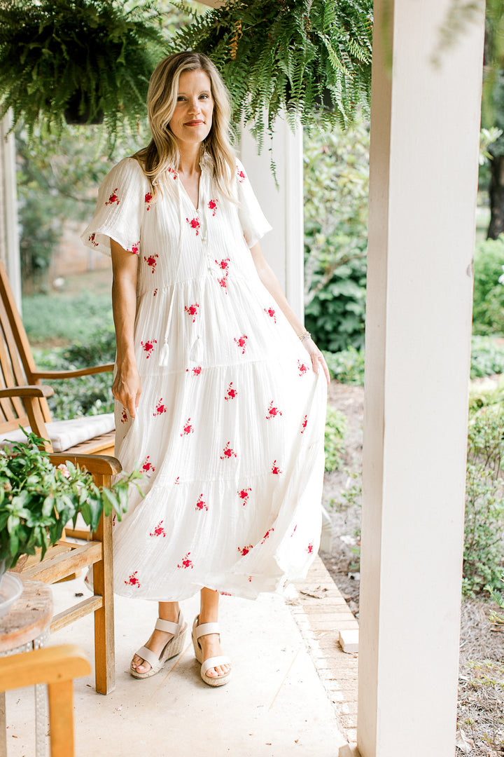 Model wearing sandals and a white maxi with red flowers, short sleeves and a v-neck.
