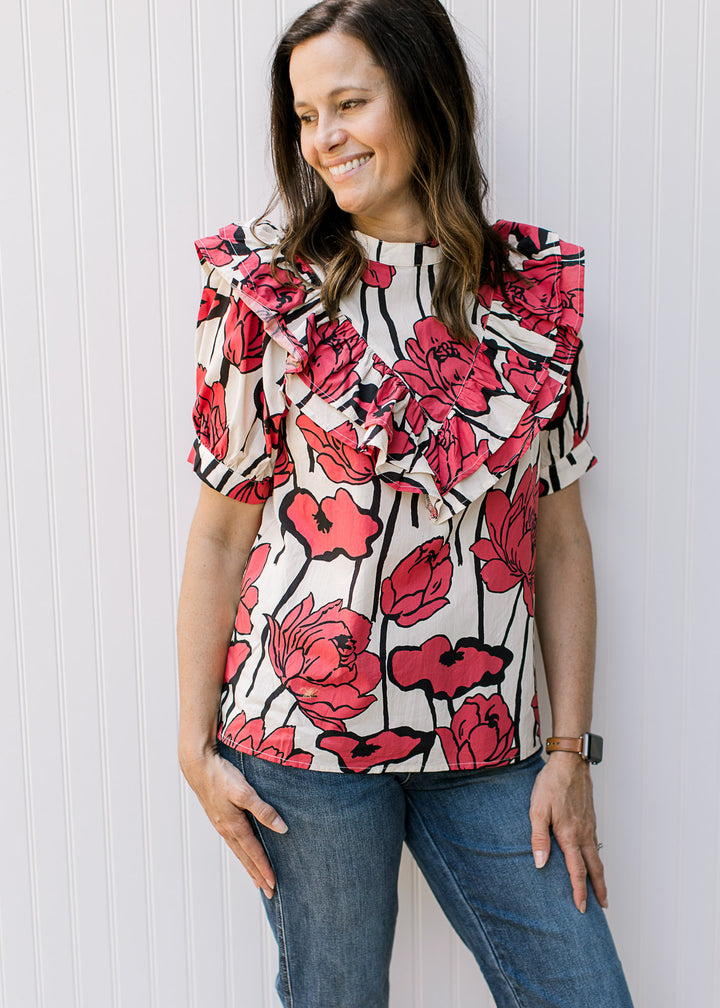 Model wearing a cream cotton top with rose colored flowers, a bib ruffle detail and short sleeves.