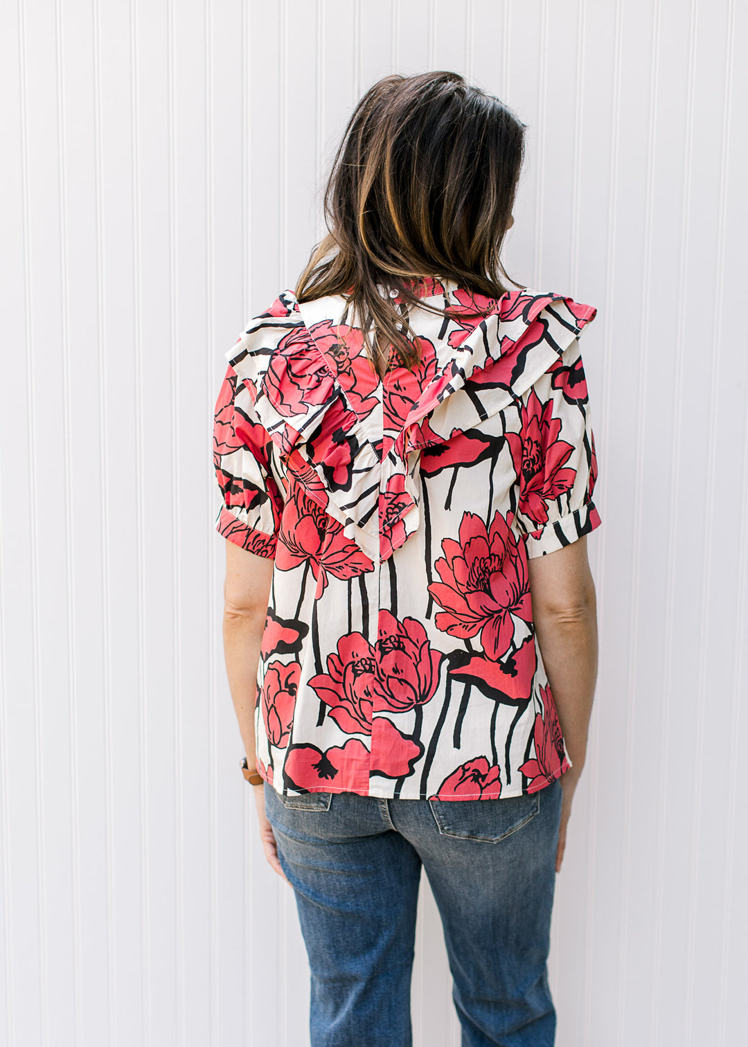 Back view of Model wearing a cream top with rose flowers, a bib ruffle detail and short sleeves.
